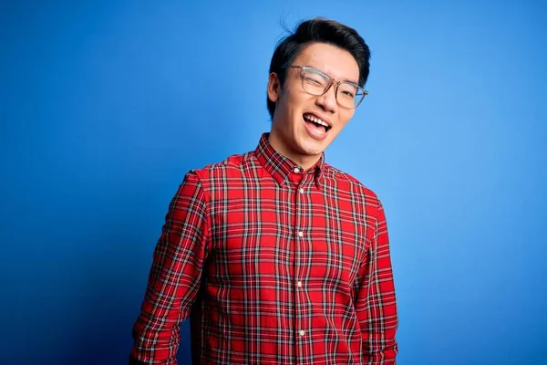 若いですハンサムな中国人男性身に着けていますカジュアルシャツと眼鏡上の青の背景ウィンク見ますザカメラとともにセクシー式 陽気で幸せな顔 — ストック写真