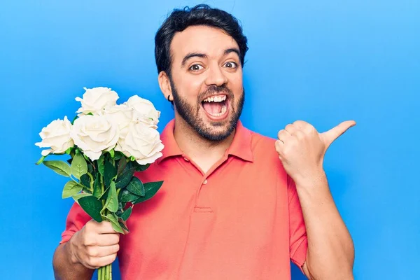 Joven Hispano Sosteniendo Flores Apuntando Con Pulgar Hacia Costado Sonriendo —  Fotos de Stock