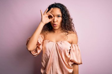 Kıvırcık saçlı, günlük tişörtünü giymiş pembe arka planda dikilmiş şaşırmış yüz ifadesiyle şaşırmış, parmaklarından bakan güzel bir kadın. İnançsız ifade.