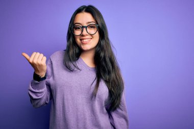Mor izole edilmiş arka planda gözlük takan genç esmer kadın gülümsüyor ve başparmağıyla yan tarafı işaret ediyor..