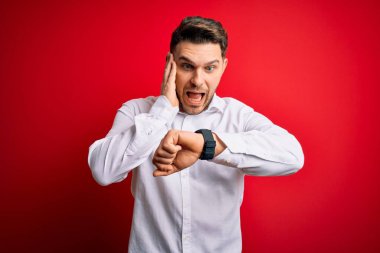 Mavi gözlü, zarif gömlekli genç bir iş adamı kırmızı izole arka planda dikiliyor. Endişeli saate bakıyor, geç kalmaktan korkuyor.