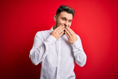 Mavi gözlü, zarif gömlekli genç bir iş adamı kırmızı izole arka planda dikiliyor gülüyor ve utanıyor kıkırdıyor elleri, dedikoduları ve skandalları ağızlarına tıkıyor.