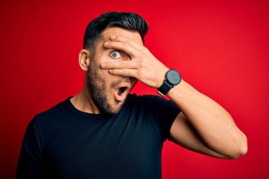 Siyah bir tişört giyen genç yakışıklı, izole edilmiş kırmızı arka planda dikiliyor. Şok olmuş bir şekilde yüzünü ve gözlerini kapıyor. Utanmış bir ifadeyle parmaklarının arasından bakıyor..