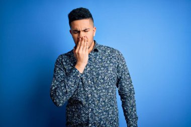 İzole edilmiş mavi arka plan üzerinde duran rahat gömlekli genç yakışıklı adam ağzıyla esnemekten sıkılmış. Huzursuz ve uykulu..
