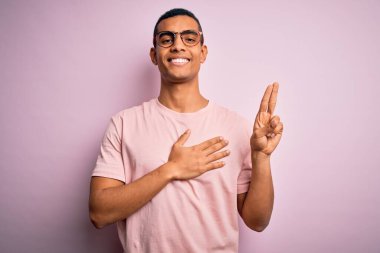 Yakışıklı Afro-Amerikalı adam pembe arka planda günlük tişört ve gözlük takıyor. Gülümsüyor, el ve göğsü havada, sadakat yemini ediyor.
