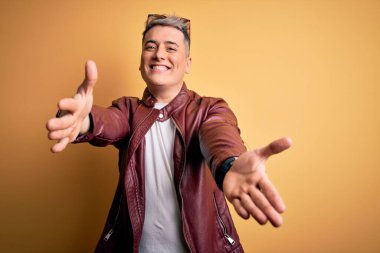 Sarı arka planda, deri ceket ve güneş gözlüğü takmış genç, yakışıklı, modern bir adam kucaklaşmak için kollarını açmış, gülümseyen kameraya bakıyor. Mutluluğu kucaklayan neşeli bir ifade..