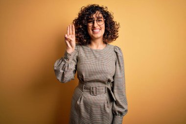 Sarı arka planda duran elbise ve gözlük takan güzel Arap iş kadını üç numaralı parmaklarıyla gülümseyerek kendinden emin ve mutlu bir şekilde işaret ediyor..