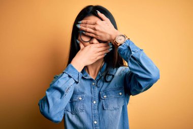 Genç, güzel, Çinli bir kadın izole edilmiş sarı arka planda sıradan bir kot gömlek giyiyor gözleri ve ağzı kapalı, şaşırmış ve şok olmuş. Duygularını gizliyor.