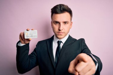 Genç beyaz iş adamı, kredi kartını pembe arka planda tutuyor. Kamerayı işaret ediyor. Ve sen, el işareti, ön taraftan pozitif ve kendinden emin bir hareket.