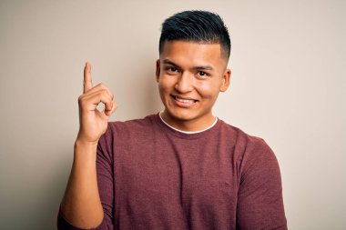 İzole edilmiş beyaz arka plan üzerinde duran gündelik süveter giyen genç, yakışıklı Latin bir adam. Yüzünde kocaman bir gülümseme, eli ve parmağı kameraya bakıyor..