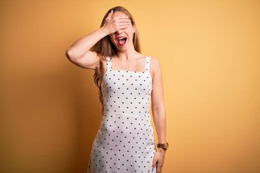 Sarı arka plan üzerine yaz elbisesi giymiş, gülümseyen ve gülen genç, güzel sarışın kadın şaşırmak için gözlerini kapatıyor. Kör kavram.