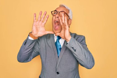 Orta yaşlı, kır saçlı, yakışıklı iş adamı sarı arka planda gözlüklü, elleri ağızında sinirli bir şekilde bağırıyor.