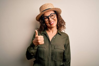 Gözlüklü ve şapkalı orta yaşlı esmer kadın izole edilmiş beyaz arka planda durup mutlu bir el hareketi yapıyor. Kameraya bakıp başarıyı gösteren ifade onaylanıyor.