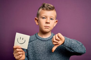 Küçük beyaz bir çocuk kağıtta gülen surat ifadesini kızgın yüzlü mutlu bir mesaj olarak gösteriyor. Negatif işaret baş parmakları aşağıda, reddedilme konsepti ise...