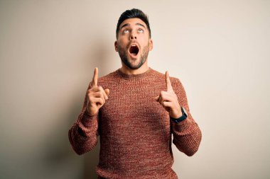 İzole edilmiş beyaz arka plan üzerinde duran gündelik süveter giyen genç yakışıklı adam şaşırmış ve şaşırmış bir şekilde yukarı bakıyor ve parmaklarıyla işaret ediyor ve kolunu kaldırıyor..