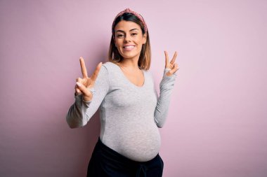Genç, güzel, esmer bir kadın hamile. Pembe arka planda bebek bekliyor. Kameraya bakıp zafer işareti yaparken gülümsüyor. İki numara..
