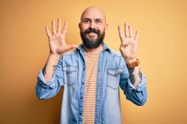 Sakallı, sıradan bir kot ceket ve çizgili tişört giyen yakışıklı kel adam dokuz numarayı gösteriyor ve gülümsüyor ve mutlu..