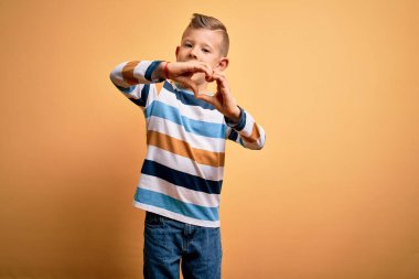 Mavi gözlü, sarı arkaplan üzerine çizgili tişört giyen, aşıkken gülümseyen, elleriyle kalp sembolü yapan küçük beyaz bir çocuk. Romantik konsept.