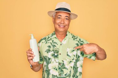 Orta yaşlı, kır saçlı, yaz şapkası takan, elinde güneş kremi tutan, sürpriz yüzlü, kendini gösteren bir adam.