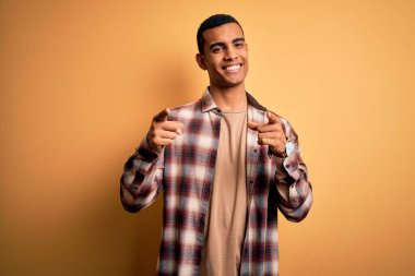 Sarı arka plan üzerinde dikilmiş, mutlu ve komik yüzlü parmakları kameraya gösteren, gündelik gömlekler giyen genç, Afro-Amerikan bir adam. İyi enerji ve titreşimler.