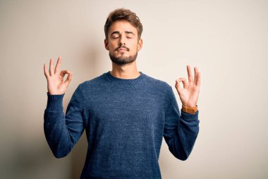Sakallı, sıradan bir süveter giyen, beyaz arka plan üzerinde duran, gözleri kapalı, parmaklarıyla meditasyon hareketi yapan genç yakışıklı bir adam. Yoga konsepti.