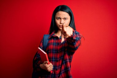 Genç, güzel Çinli bir öğrenci elinde kitapla pembe arka planda dikilip kamerayı işaret ediyor ve sen, el işareti, ön taraftan pozitif ve kendinden emin bir jest.