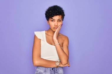 Genç ve güzel Afro Amerikalı kadın mor arka plan üzerine günlük tişört giyiyor. Kollarını kavuşturup depresyondan sıkılmış ve yorgun görünüyor..