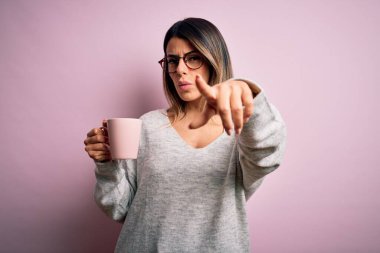 Genç, güzel, esmer, gözlüklü bir kadın pembe kahve fincanı içerek kamerayı işaret ediyor ve sen, el işareti, ön taraftan pozitif ve kendinden emin bir jest.