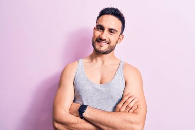 Sakallı, pembe arka planda kolsuz tişört giyen, kollarını kavuşturup kameraya bakan yakışıklı genç bir adam. Pozitif kişi.