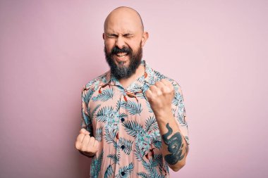 Sakallı ve dövmeli yakışıklı bir adam pembe arka planda sıradan bir çiçek gömleği giyiyor. Şaşırmış ve şaşırmış bir şekilde kolları havada ve gözleri kapalı bir şekilde başarıyı kutluyor. Kazanan konsept.