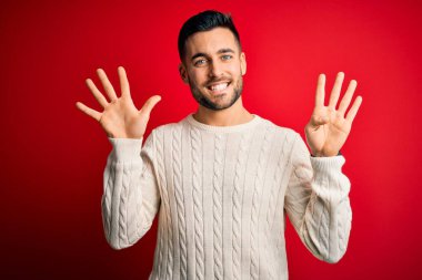 İzole edilmiş kırmızı arka planda duran beyaz süveter giyen genç ve yakışıklı bir adam dokuz numaralı parmaklarıyla işaret ederken kendinden emin ve mutlu bir şekilde gülümsüyor..