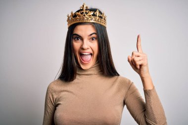 İzole edilmiş beyaz arka plan üzerinde altın kraliçe tacı takan genç, güzel esmer kadın başarılı bir fikir işaret ediyor. Heyecanlı ve mutlu. Bir numara..