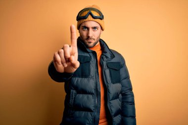 Kar giysileri ve kayak gözlüğü takan sakallı genç yakışıklı kayakçı parmağını kaldırmış ve kızgın bir ifade sergiliyor.