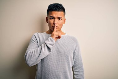 Genç, yakışıklı, latin bir adam, günlük süveter giyip izole edilmiş beyaz arka planda dikiliyor ve parmakları dudaklarında sessiz olmak istiyor. Sessizlik ve gizli konsept.