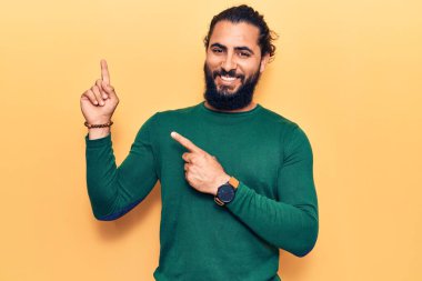 Gündelik kıyafetler giyen genç Arap adam gülümseyip kameraya bakıyor ve iki eliyle ve parmaklarıyla kenarı işaret ediyor.. 