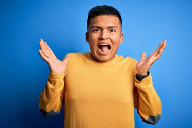 Sarı günlük kazak giyen genç, yakışıklı latin adam mavi arka planda çılgınca kutlama yapıyor ve kollarını kaldırıp heyecanla haykırarak başarıyı kutluyor. Kazanan konsept