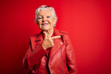 Son sınıf güzeli, gri saçlı, günlük kırmızı ceket ve güneş gözlüğü takan neşeli, yüzünde mutlu ve doğal bir ifadeyle el ve parmaklarıyla işaret eden bir gülümsemesi olan bir kadın.