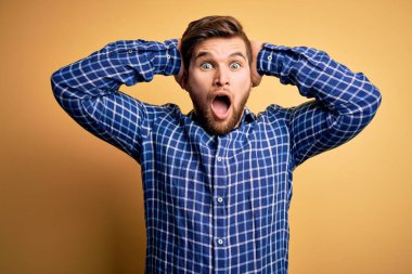 Sarışın, sakallı ve mavi gözlü genç bir işadamı sarı arka planda tişört giyiyor ve elleri başında korkmuş, korkmuş ve ağzı açık şoktan şaşırmış.