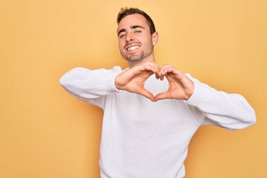 Mavi gözlü, sıradan bir süveter giyen, sarı arka planda dikilmiş, aşık olmuş, elleriyle kalp sembolü yapan genç yakışıklı bir adam. Romantik konsept.