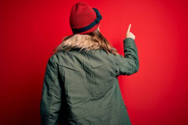 Kayak gözlüğü takan ve kırmızı arka planda kayak yapmak için kışlık ceket giyen genç sarışın kız parmağıyla öne doğru poz veriyor.