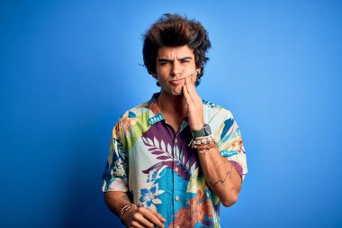 İzole edilmiş mavi arka plan üzerinde tatilde olan genç yakışıklı bir adam diş ağrısı ya da diş ağrısı yüzünden ağız ve acı dolu bir ifadeyle dokunuyor. Dişçi