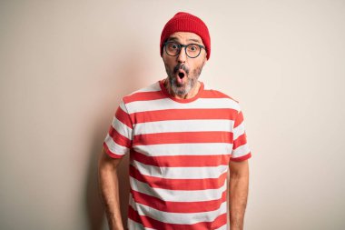 Orta yaşlı, beyaz tişört gözlüklü, beyaz arka plan kaplamalı beyaz bir adam korku, korku ve heyecan dolu bir yüz ifadesinden korkuyor ve şoka uğruyor..