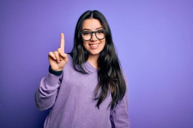 Genç esmer kadın mor renkli gözlük takıyor. İzole edilmiş arka planda. Bir numarayı işaret ediyor ve gülümsüyor..