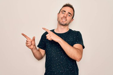 Mavi gözlü, sıradan tişörtlü, beyaz arka planda duran, gülümseyen ve kameraya bakan, iki eli ve parmağı yanda olan genç, yakışıklı bir adam..