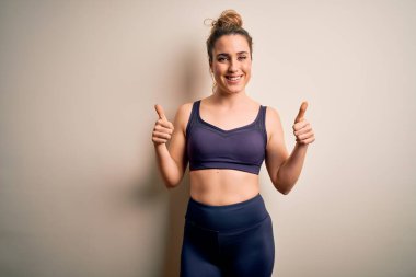 Genç, sarışın, spor yapan, spor giyinen, beyaz arka plan başarısı tabelasına spor giyinen, el hareketleriyle olumlu jest yapan, gülümseyen ve mutlu bir kadın. Neşeli ifade ve kazanan jest.