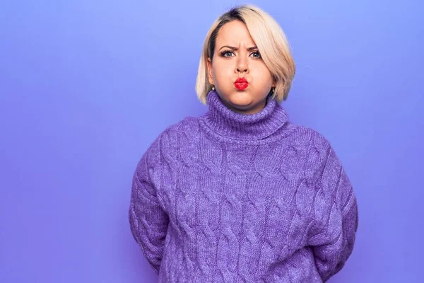 Güzel sarışın ve cüsseli bir kadın mor arka planda komik yüzlü şişen yanakların üzerine balıkçı yaka kazak giyiyor. Havayla şişmiş ağız, çılgın bir ifade..