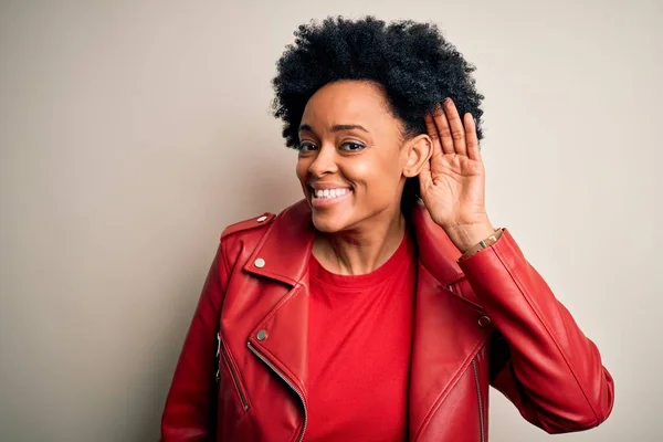 Young beautiful African American afro woman with curly hair wearing casual red jacket smiling with hand over ear listening an hearing to rumor or gossip. Deafness concept.