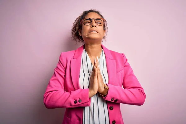 Beautiful african american businesswoman wearing jacket and glasses over pink background begging and praying with hands together with hope expression on face very emotional and worried. Begging.