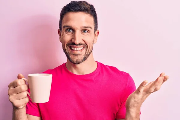 Young handsome man drinking cup off coffee standing over isolated pink background celebrating achievement with happy smile and winner expression with raised hand