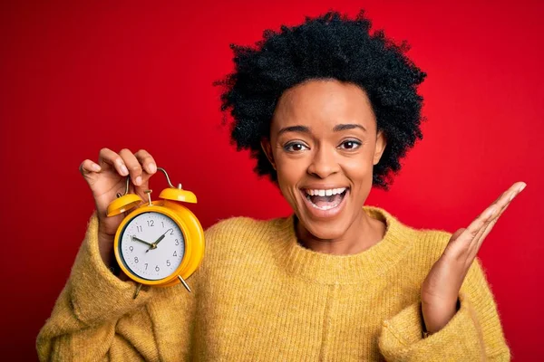 Young African American afro woman with curly hair holding vintage alarm clock very happy and excited, winner expression celebrating victory screaming with big smile and raised hands